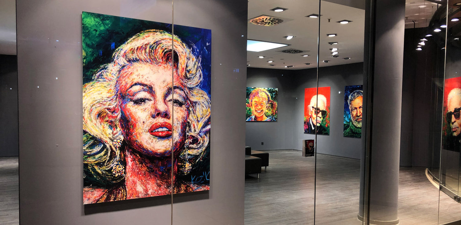View through the gallery window showing Marilyn Monroe’s empathic portrait by Marcel Kozik, displayed alongside several colorful expressive portraits on the walls — modern art exhibition with vibrant lighting.