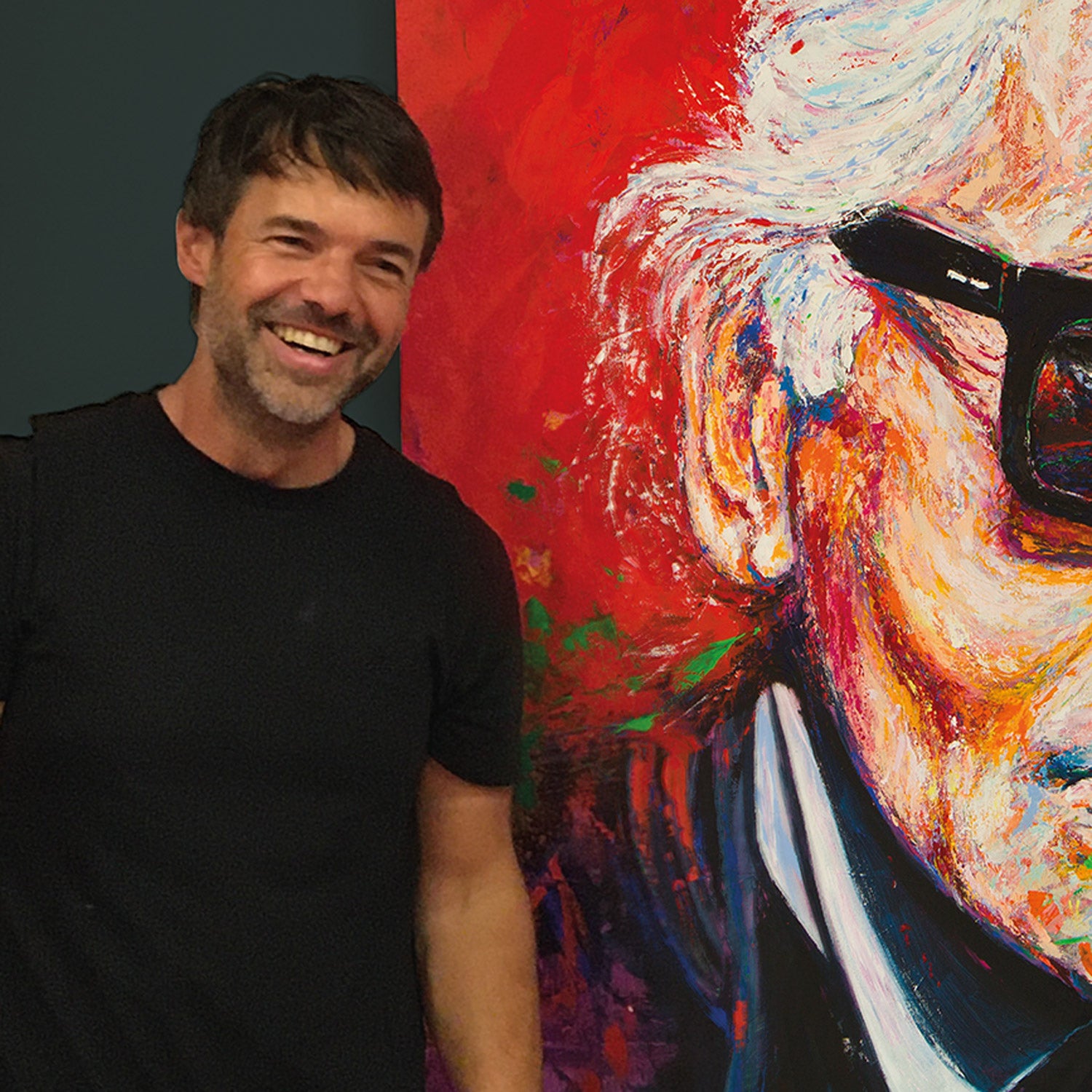 The artist standing beside a large expressive acrylic painting inspired by Karl Lagerfeld, part of the Empathism art movement. The 120×160 cm portrait features vivid layers of color that symbolize emotional depth and life phases, reflecting the artist’s signature technique of building images like growing life rings.