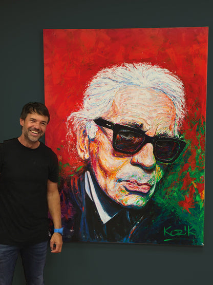 Artist Marcel Kozik standing beside his original Karl Lagerfeld empathic portrait, a 120×160 cm expressive acrylic painting with bold red and green textured strokes.